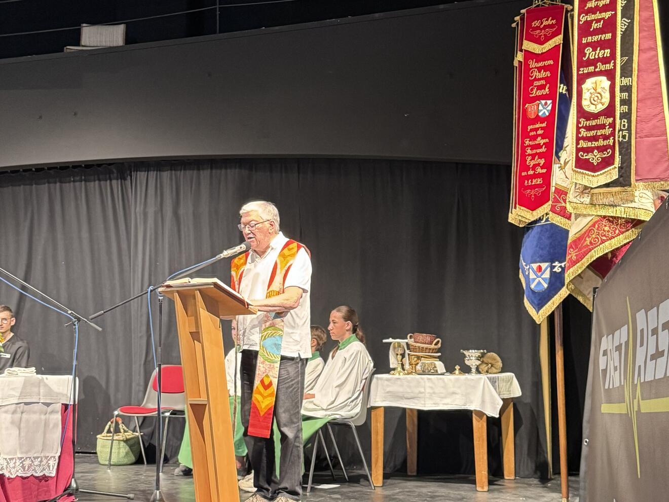 Pfarrer Kahnert zelebriert den Gottesdienst im Rahmen des Brunnenfestes im Theaterstadel Heinrichshofen.  Ministranten sind im Hintergrund. Seitlich sind die Fahen der Vereine.